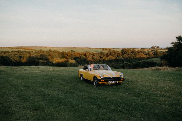 A yellow vintage wedding car parked in a green field with trees and hills beyond