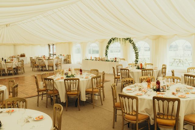 Inside the marquee: draped ivory ceiling, gold chairs at round tables and a greenery arch behind the top table