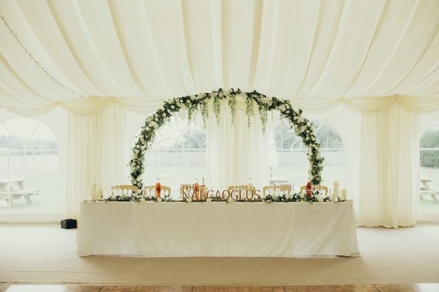 Top table in front of a round greenery and white flower arch inside the marquee