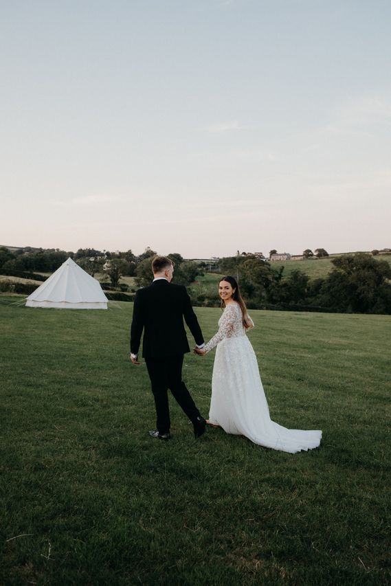 Bride and groom walking hand in hand across the fields at dusk, with a bell tent behind