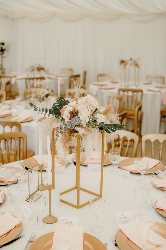 A wedding table with a tall gold flower stand, candles and blush napkins