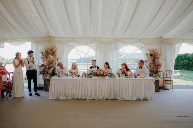 Top table dressed with pampas grass and flowers beneath the draped marquee ceiling and arched windows